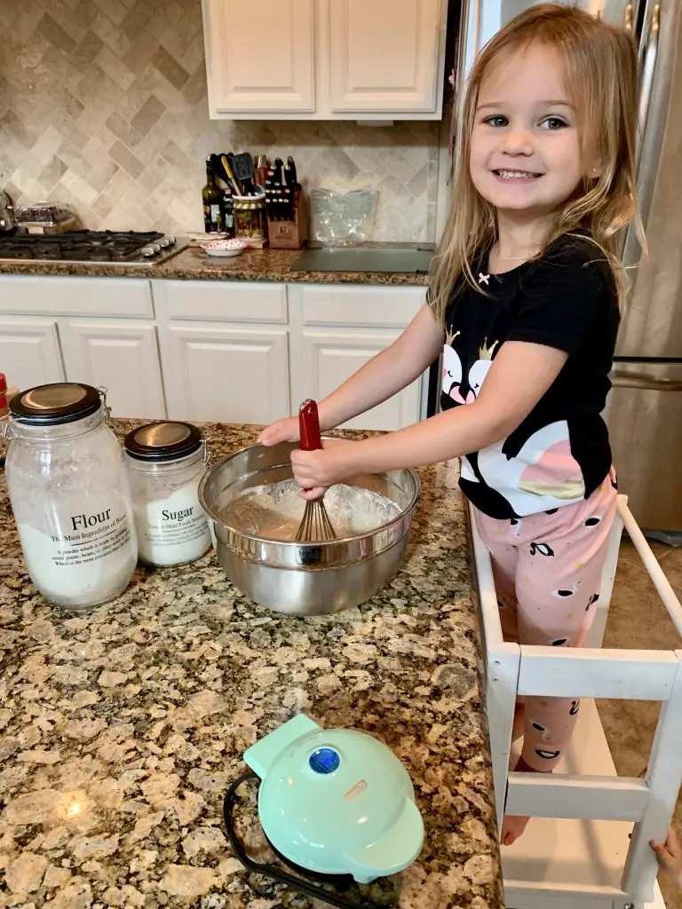 Toddler cooking waffles with mom. Flour and sugar jars. Mini waffle maker.